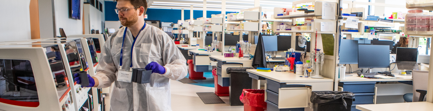 A scientist wearing a white lab coat and proper protective equipment in a fully-stocked lab