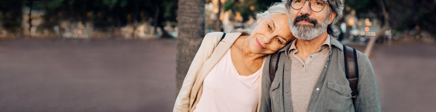 Older man standing and older woman resting her head against older mans shoulder with a tree in the background