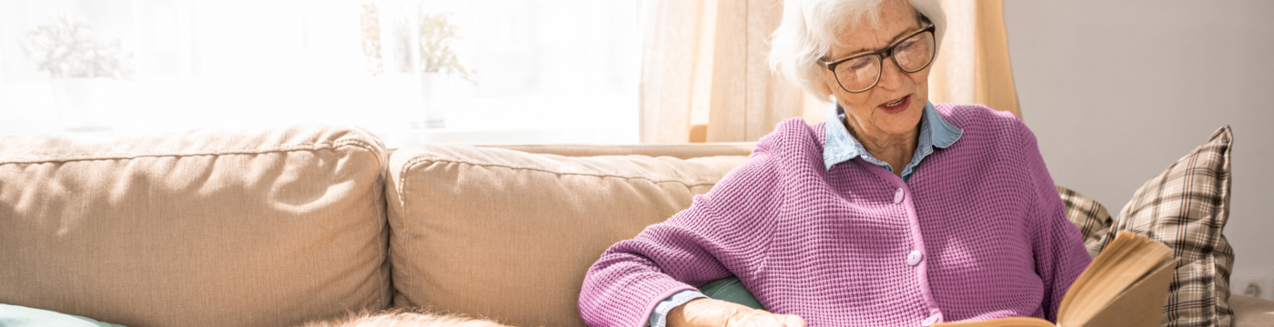 Older woman with glasses reading a book on a couch.