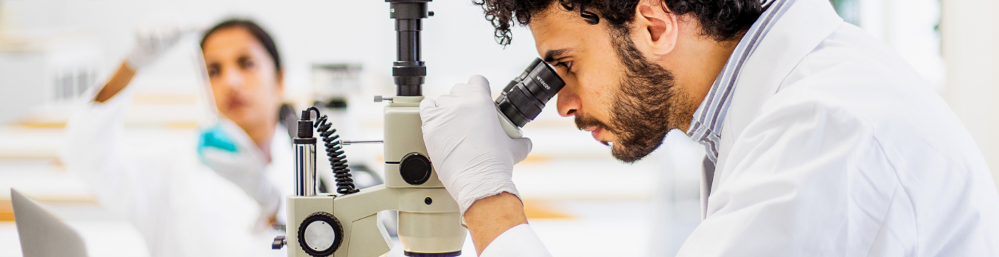 A lab technician in proper protective equipment is looking through a microscope, analyzing a slide